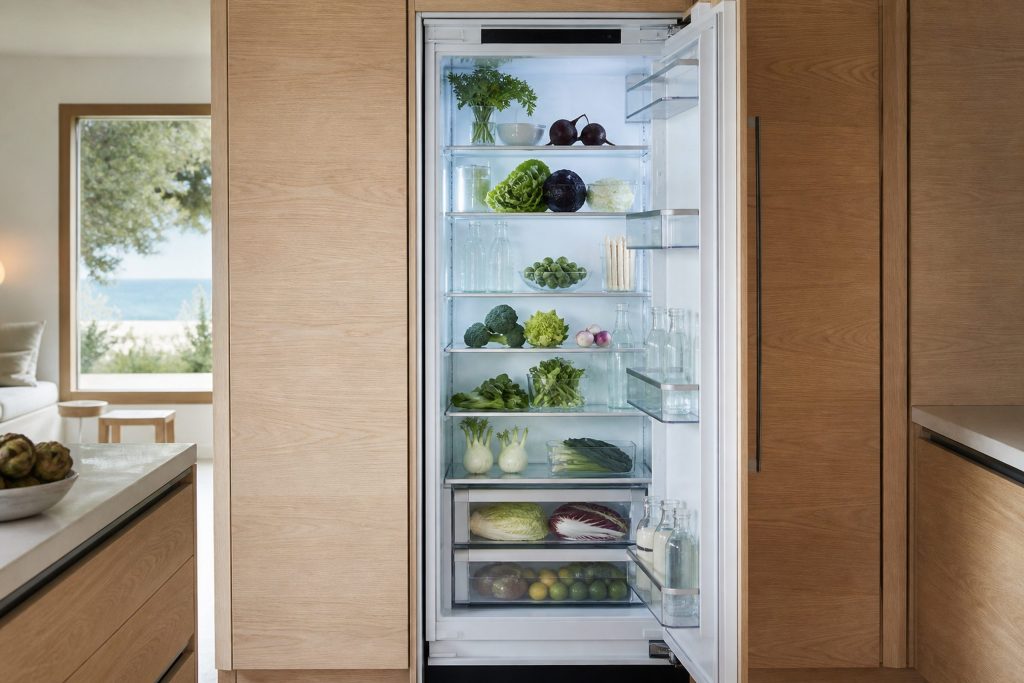 A tall integrated refrigerator column standing open in a bright coastal kitchen, glass shelves neatly filled with vegetables and glass bottles, cool even interior light, pale oak cabinetry either side.