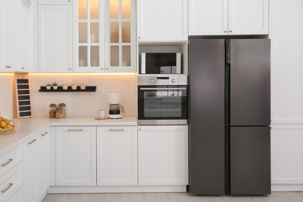 A four-door refrigerator freezer standing in a white shaker kitchen, its doors closed, daylight from the room behind