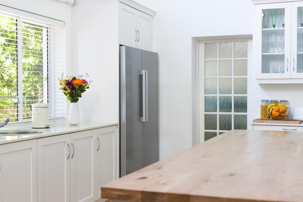 A tall freestanding refrigerator fitted into a niche in a bright white kitchen, a window and a timber worktop beside it