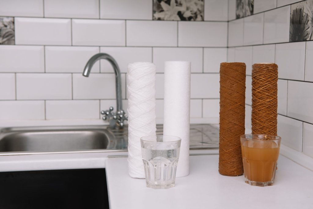 Two new white filter cartridges beside two brown exhausted ones, with a glass of clear water and a glass of dirty water