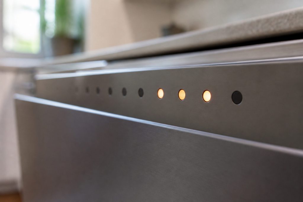 A close raking view along the top edge of a dish drawer front where a row of small round indicator lamps sits, three of them lit, no digits and no text anywhere on the strip, shallow focus.