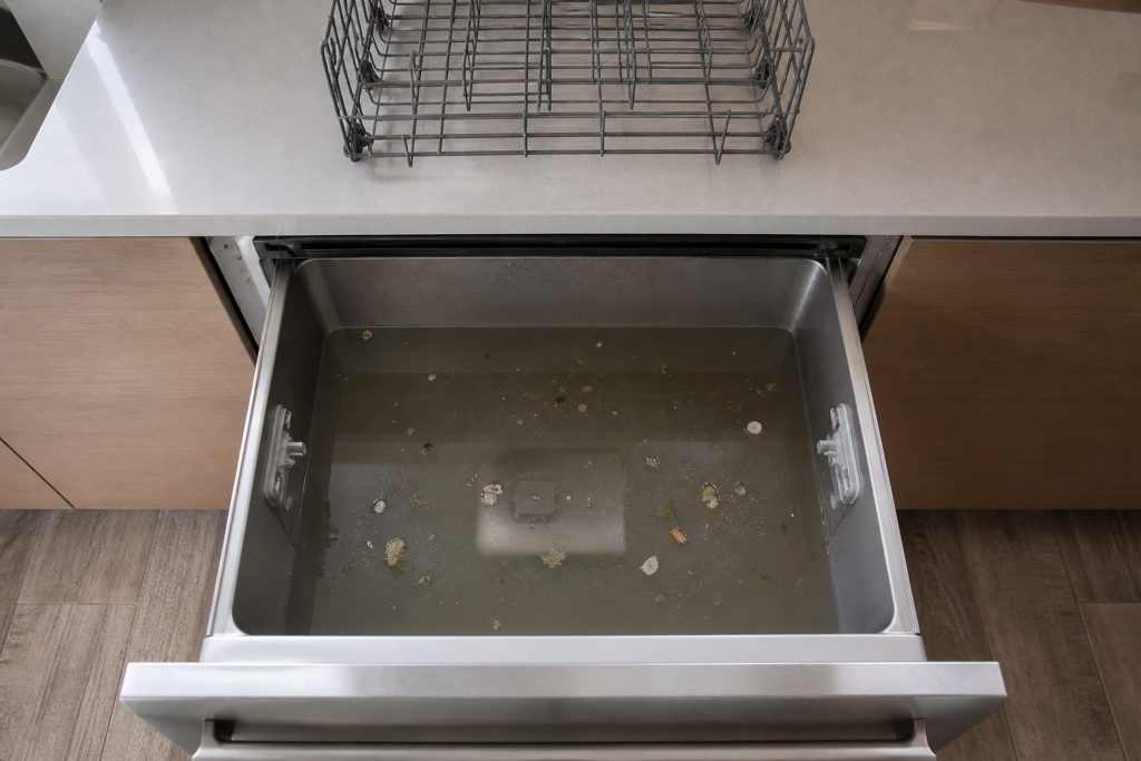 Looking down into the open tub of a dish drawer dishwasher with dirty water standing over the flat filter plate, food debris floating on it and the wire basket lifted out onto the counter