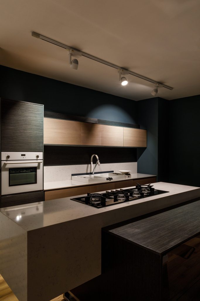 A dark blue kitchen at night, a gas hob set into a pale stone island, a white oven column at the left.