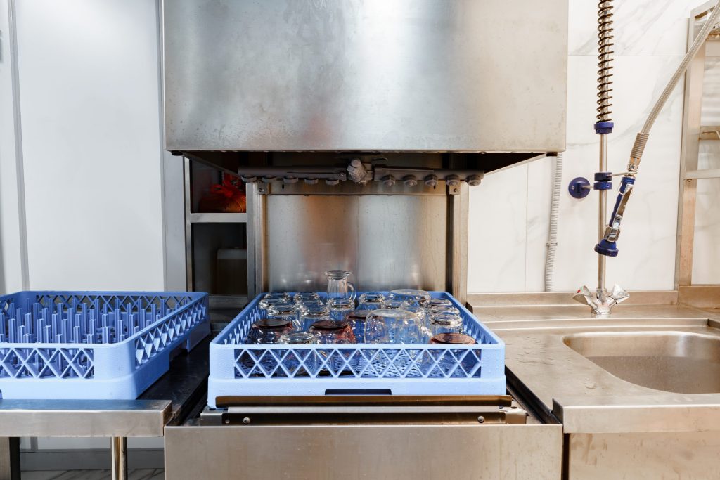 A hood-type dishwasher with its hood raised over a rack of glasses, a second rack waiting at the left.
