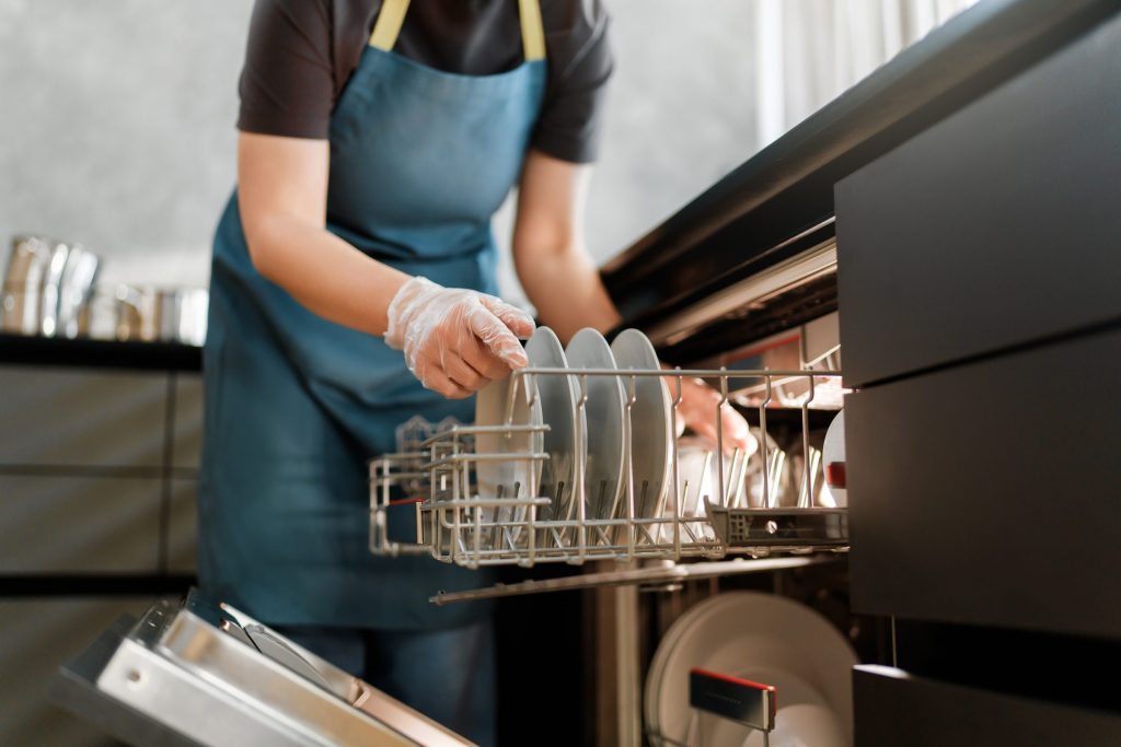 A kitchen worker in an apron loading plates into the rack of a built-under dishwasher behind a dark front.