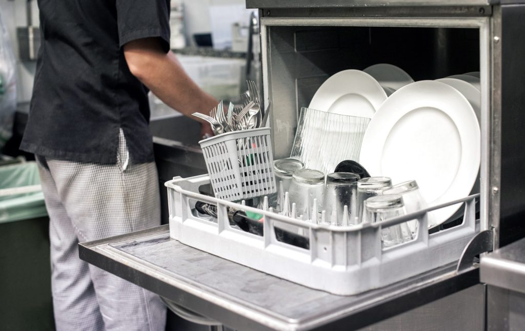 A kitchen porter sliding a loaded rack of plates and glasses into a stainless commercial dishwasher.