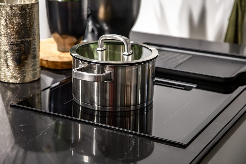 A steel pan standing on a black glass cooktop in a dark kitchen.