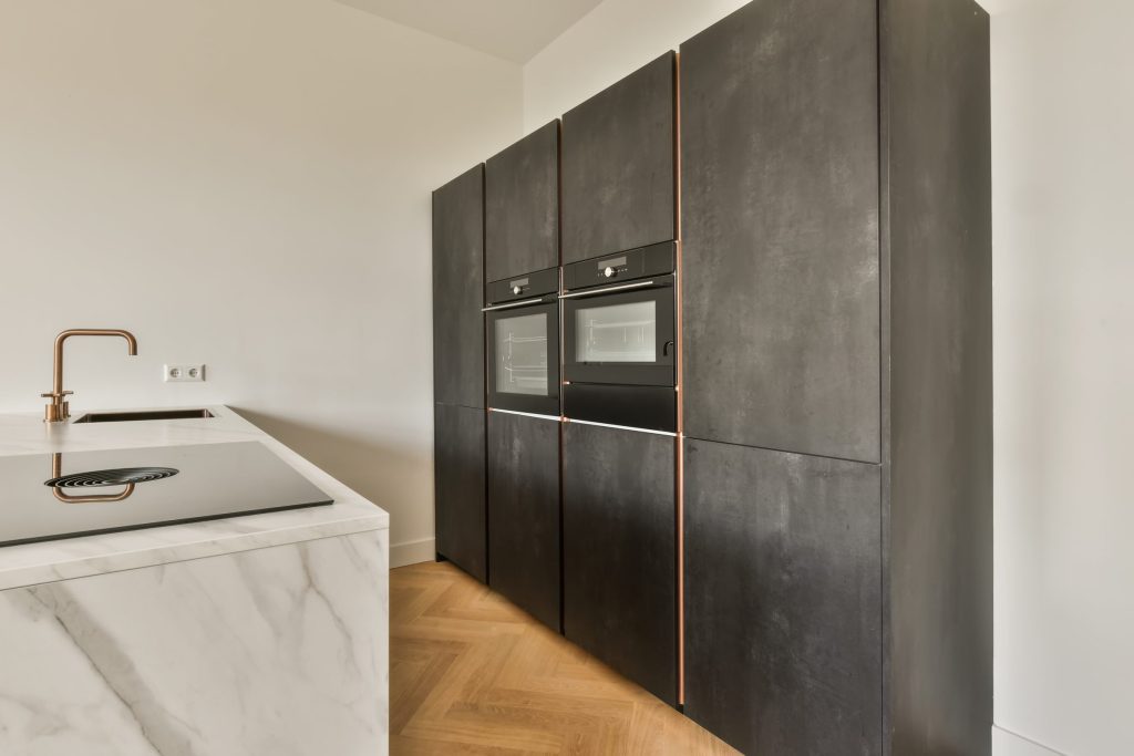 A dark tall cabinet run with two built-in ovens, copper reveals, marble island.
