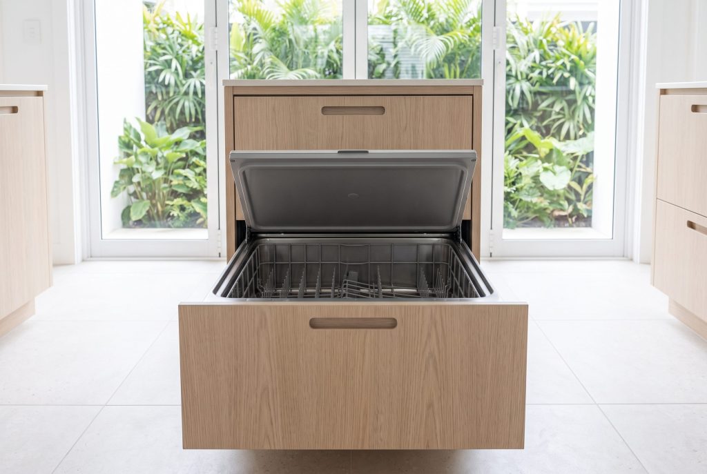 A double dish drawer dishwasher with pale oak cabinet fronts, the upper drawer pulled open and its flat lid raised clear of the empty tub, green foliage through the windows behind