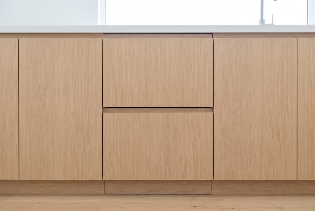 A run of pale oak cabinet fronts in which a double dish drawer dishwasher stands closed, given away only by two thin shadow gaps and a recessed finger pull