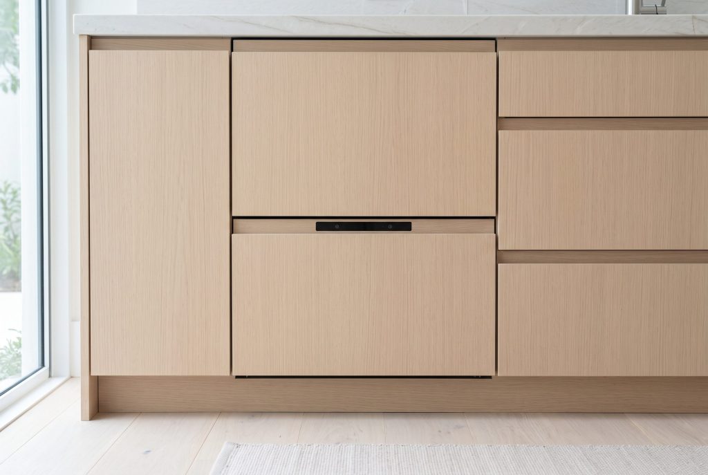 A run of pale oak cabinet fronts in which a double dish drawer dishwasher is hidden, identifiable only by two thin shadow gaps