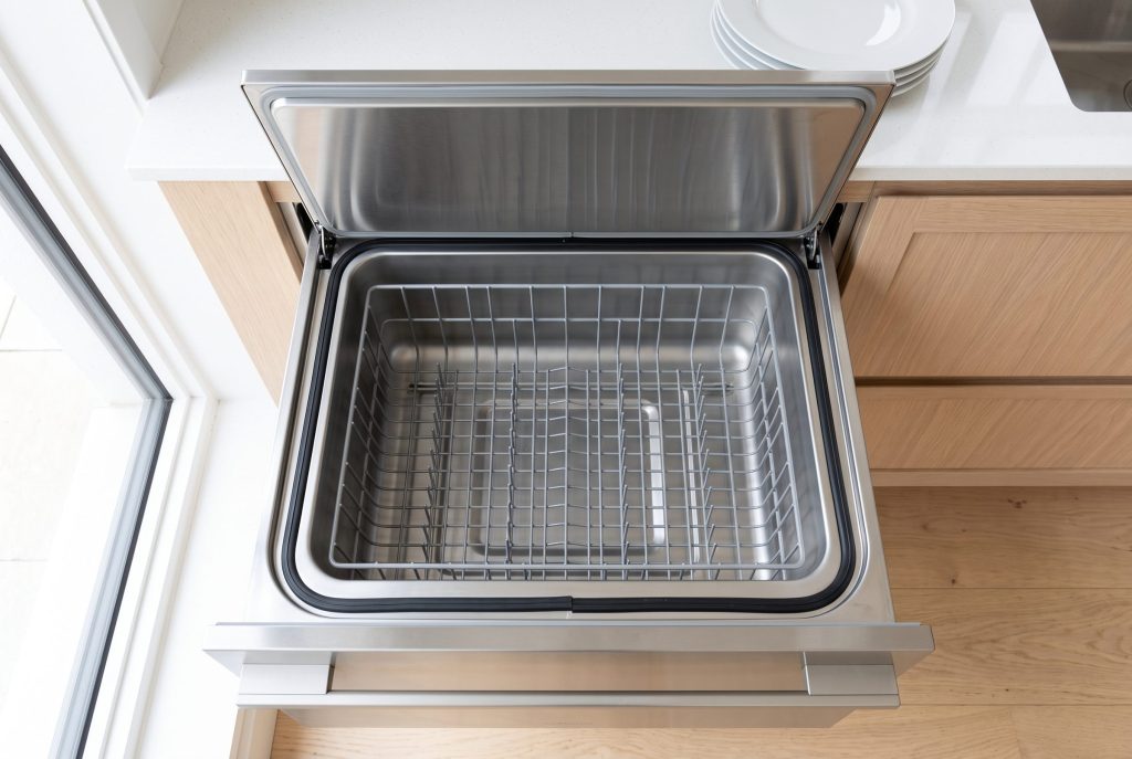 Looking down into the open tub of a dish drawer dishwasher, the flat lid raised clear above it and the wire basket empty