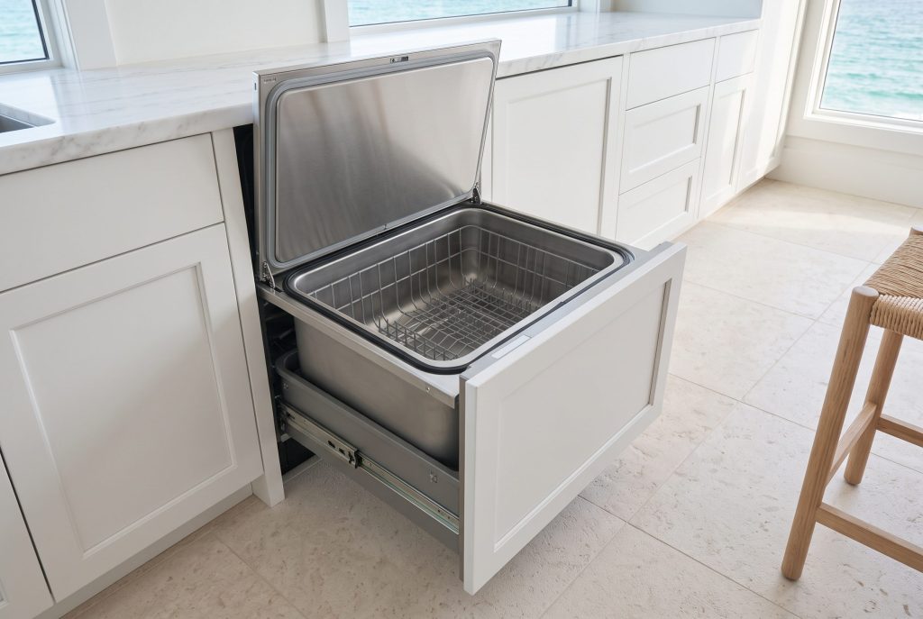 A dish drawer dishwasher open in a white kitchen, the lid raised clear of the empty stainless tub, a stool at the frame edge