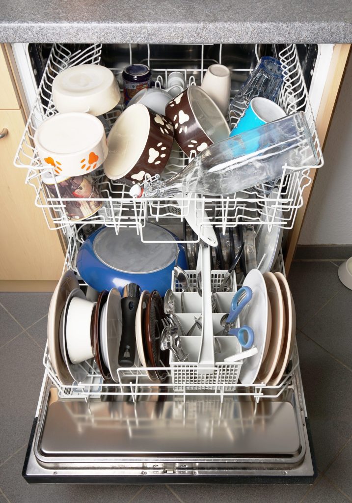 A dishwasher door down with both racks loaded — plates on edge below, cups above.