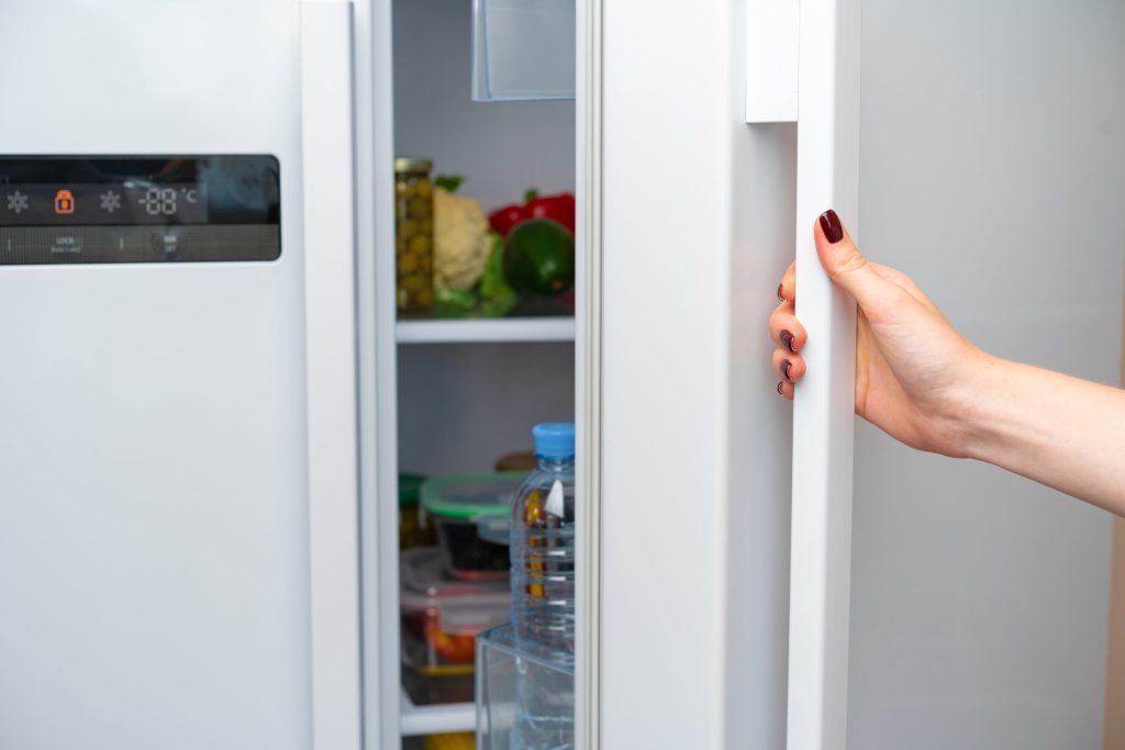 A hand pulling open the door of a side-by-side refrigerator, the control panel lit and the shelves stocked behind it.