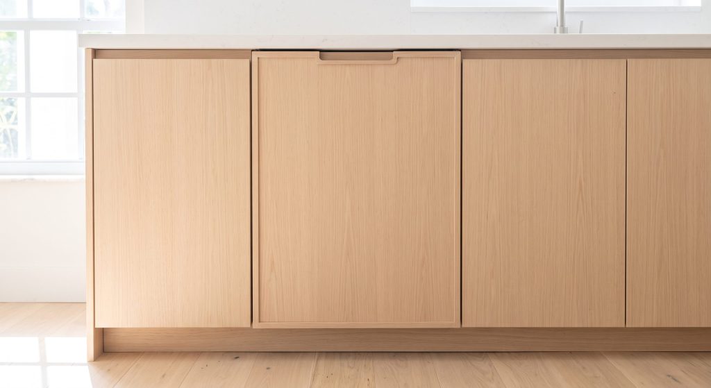 A run of pale oak cabinet fronts with a recessed finger pull, behind which an integrated refrigerator column stands closed, bright daylight from a window at the left