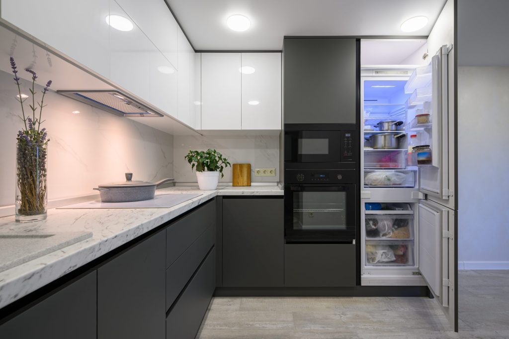 A grey and white kitchen with an oven and microwave stack, and a refrigerator standing open at the end of the run.