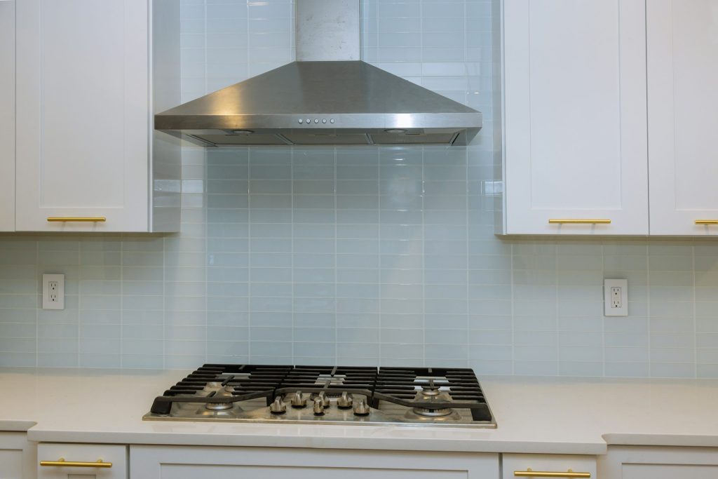 A stainless chimney hood mounted on a tiled wall above a gas cooktop in a white kitchen