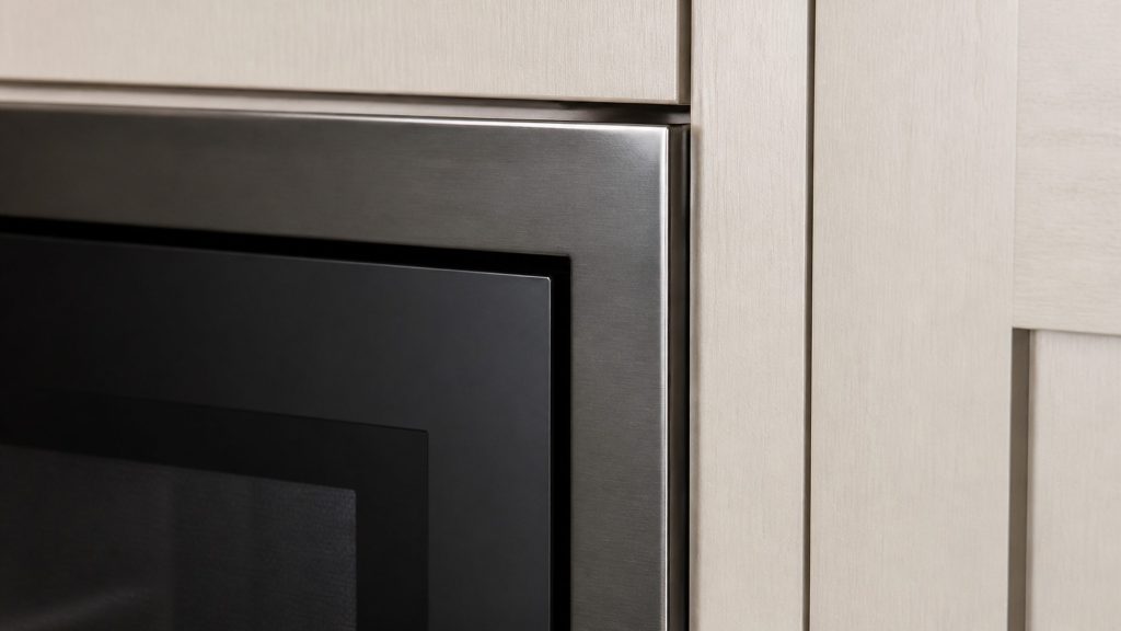 The trim kit edge where a built-in microwave meets the cabinet opening, photographed close along the frame so the joint and the door seam read.