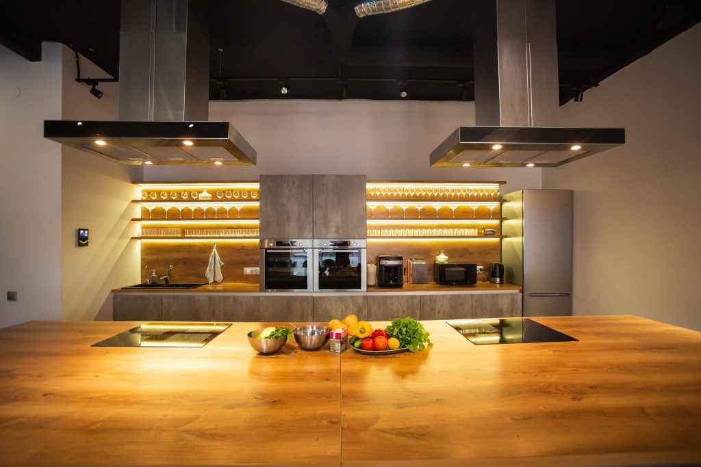 A wide oak island with two induction hobs under two stainless island hoods, glassware lit on shelves behind.