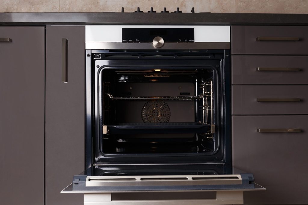 A built-in oven with a black glass fascia standing open below a gas hob in a run of dark brown cabinetry, the racks empty
