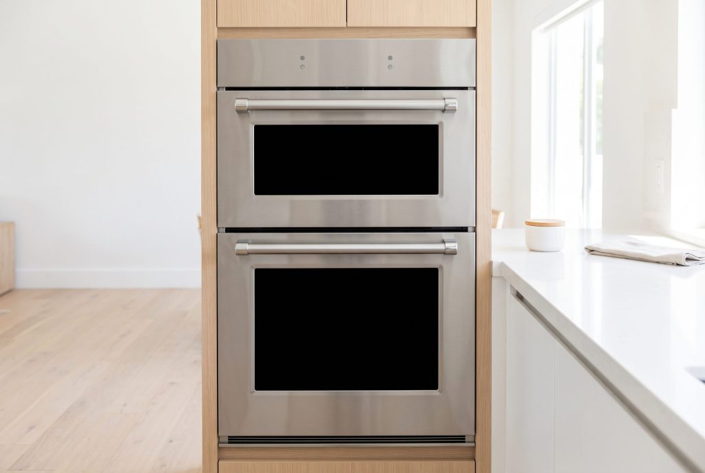 A stainless double wall oven with two black glass door panels and a heavy bar handle between them, both doors closed, white stone worktop beside it
