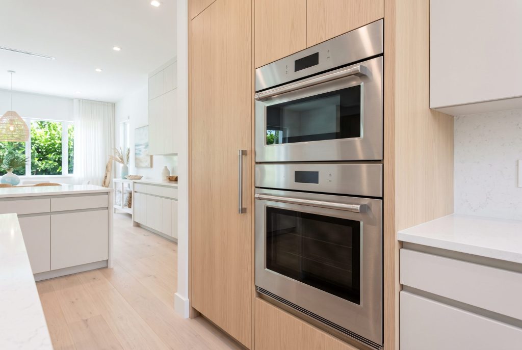 A stainless double wall oven set in a tall pale oak column, both doors closed, seen from a few steps back across a bright open-plan kitchen