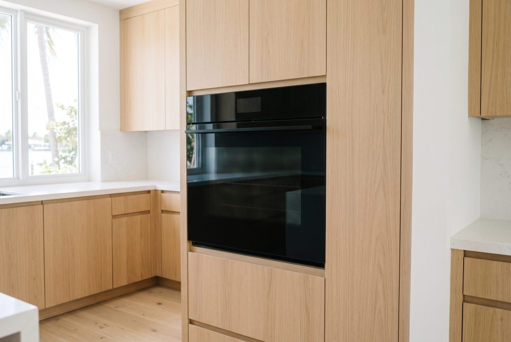 A built-in wall oven with a flush handleless front of black glass set into a tall pale oak cabinet column, the glass clean and reflecting a bright coastal kitchen with water through the window