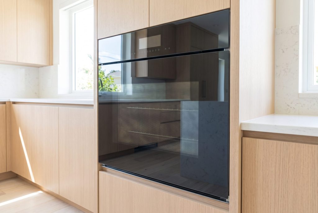 A compact wall oven with a flush black glass front set into a pale oak column, door closed, a bright coastal kitchen and the sea reflected in the glass