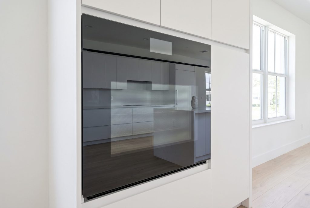 A handleless wall oven with a dark glass front set into a white run between two windows, the door closed and the room reflected in the glass