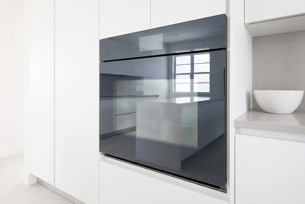 A wall oven with a flush dark glass front in a white corner run, door closed, a white bowl on the worktop beside it
