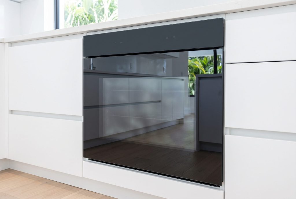 A wall oven with a flush front of dark glass set low into a white handleless kitchen run, its door closed and the glass reflecting the room