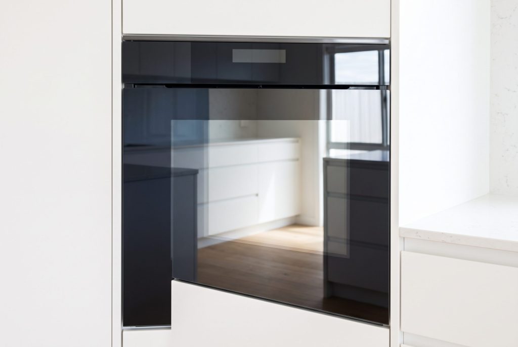 A wall oven with a flush dark glass front set in a white cabinet column, door closed, cool daylight across the glass