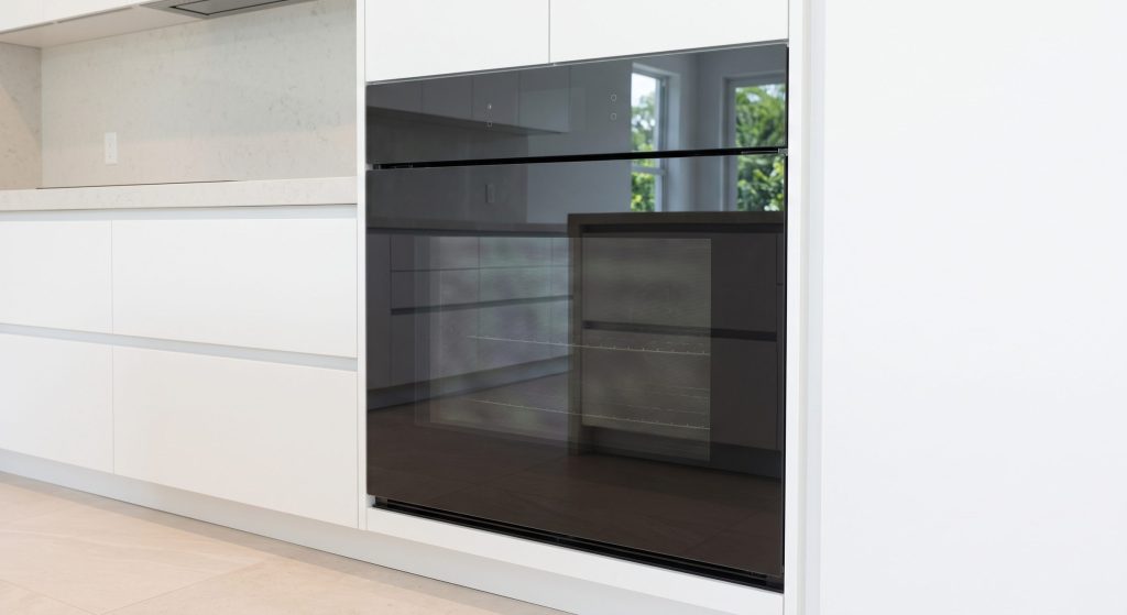 A wall oven with a flush handleless front of dark glass in a white handleless kitchen, the glass clean and reflecting the room