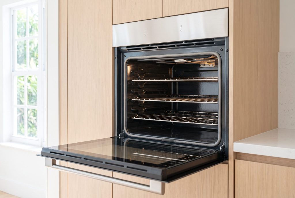 A wall oven standing open in a pale oak column, the drop-down door out flat and four empty racks lit by the cavity lamp