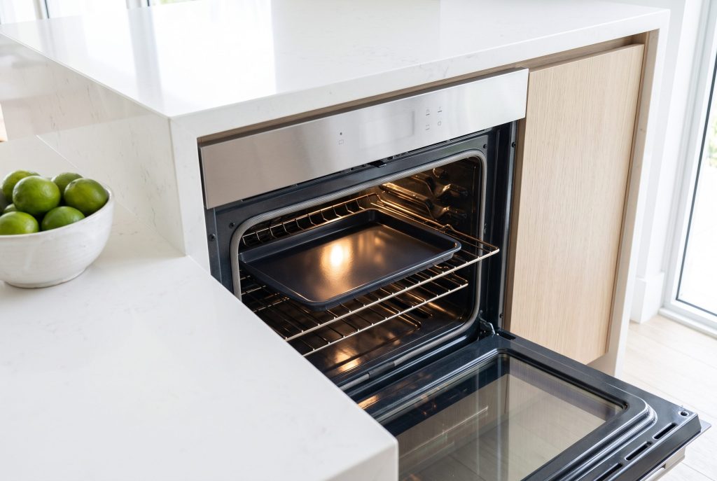 A wall oven standing open with a single empty baking tray on the middle rack under the cavity lamp, a bowl of limes on the stone worktop beside it