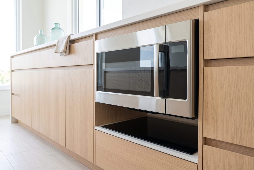 An over-the-range microwave oven mounted above a flush cooktop in a pale oak kitchen run, its door closed