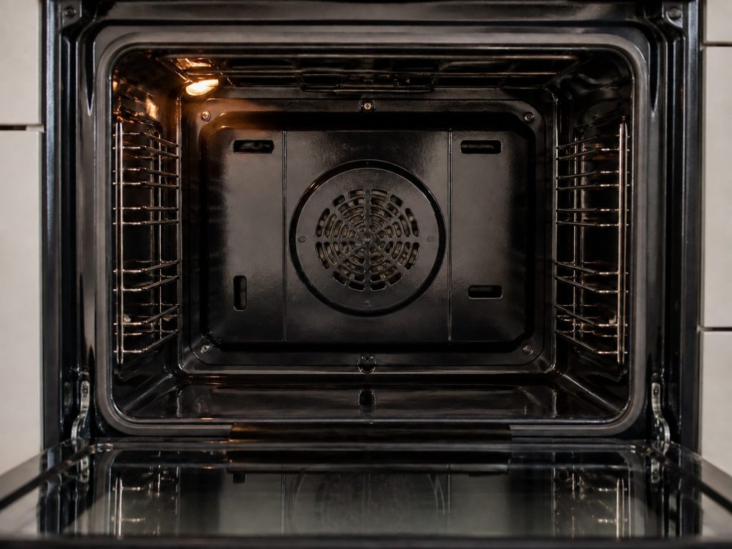 The back wall of an oven cavity with the convection fan cover and its circular grille in the center, the door open and the cavity light on.