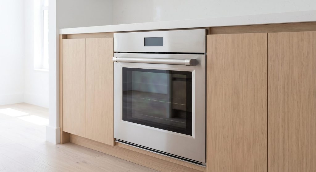A single stainless wall oven built low into a pale oak run, its door closed and the glass reflecting the bright room