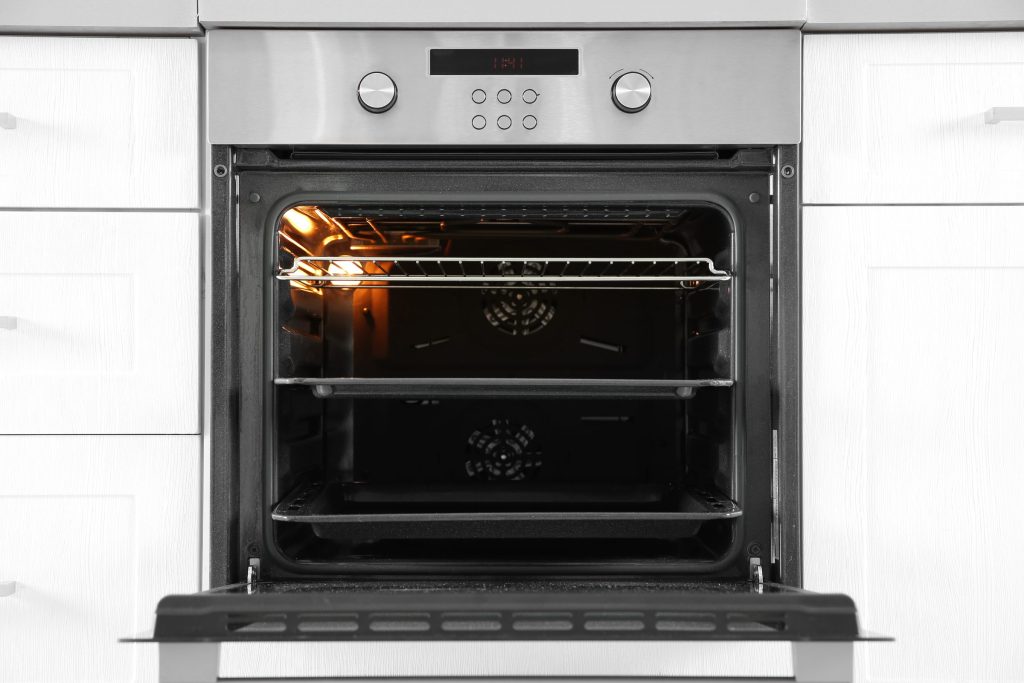 A stainless built-in oven standing open in a white kitchen, the cavity lit and the racks empty, two round control dials on the fascia above