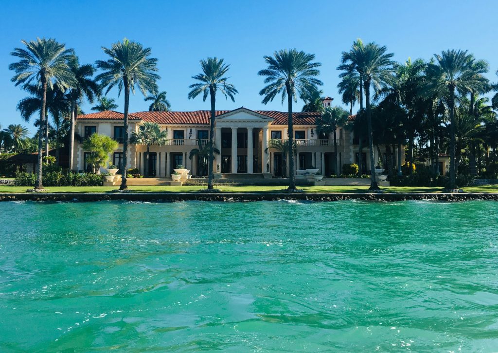 A large waterfront house behind a row of palms, seen across the water