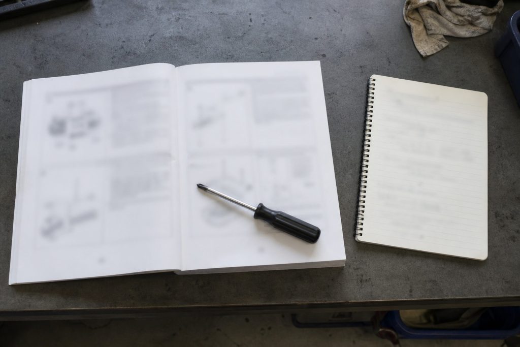A technical manual open flat on a work surface with a spiral notepad beside it, both blurred past legibility, a small screwdriver acting as a page weight, cool overhead daylight.