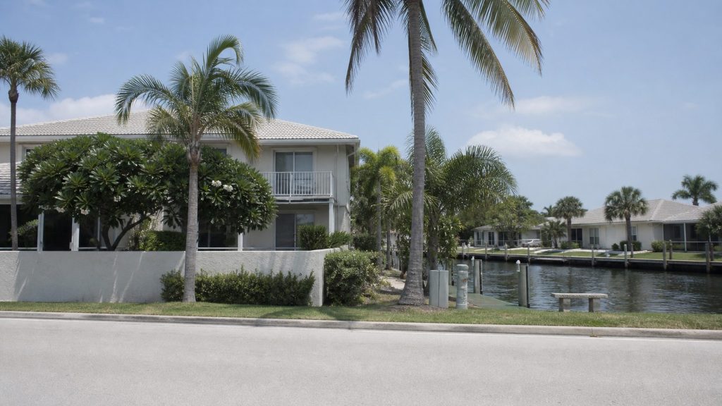 A low-rise waterfront residential street in South Florida in flat bright daylight — pale stucco walls, palms, a canal edge — photographed from across the road with no signage and no recognisable landmark.