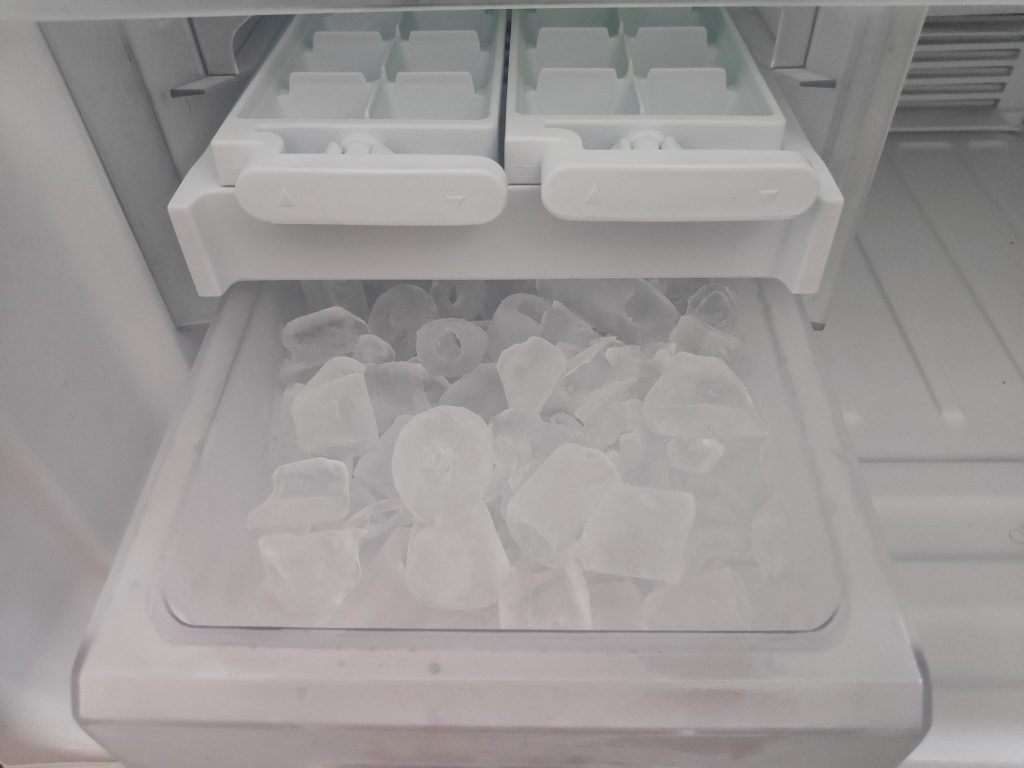 An ice maker bin filled with cubes below the molds, seen from above inside a white freezer