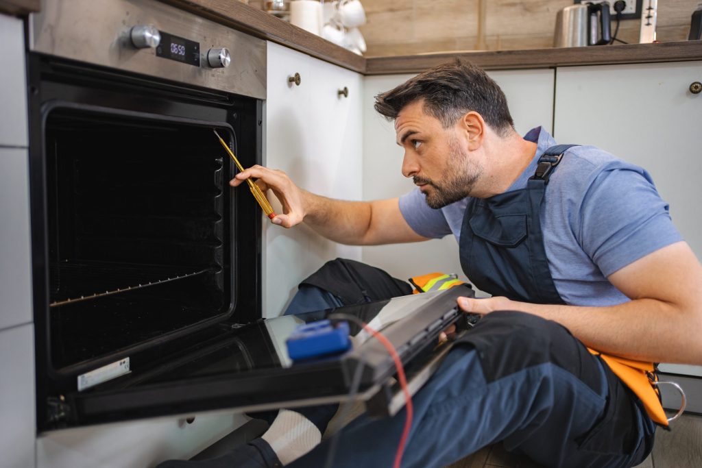 A technician kneeling at an open built-in oven with a multimeter, probes on the cavity wiring.