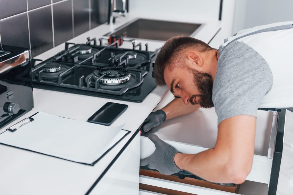 A technician leaning under a gas cooktop to check the cabinet below.
