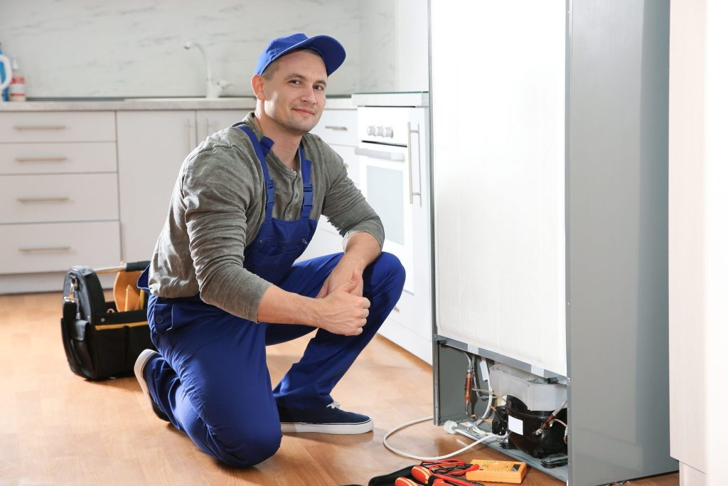 A technician kneeling at the base of a tall refrigerator with the lower panel off