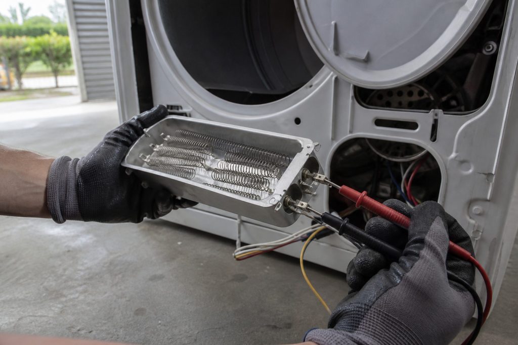 A dryer heating element assembly withdrawn from the back of the machine and held out on its housing in one gloved hand, the coil clearly visible inside it, meter probes from the other hand resting on its terminals.
