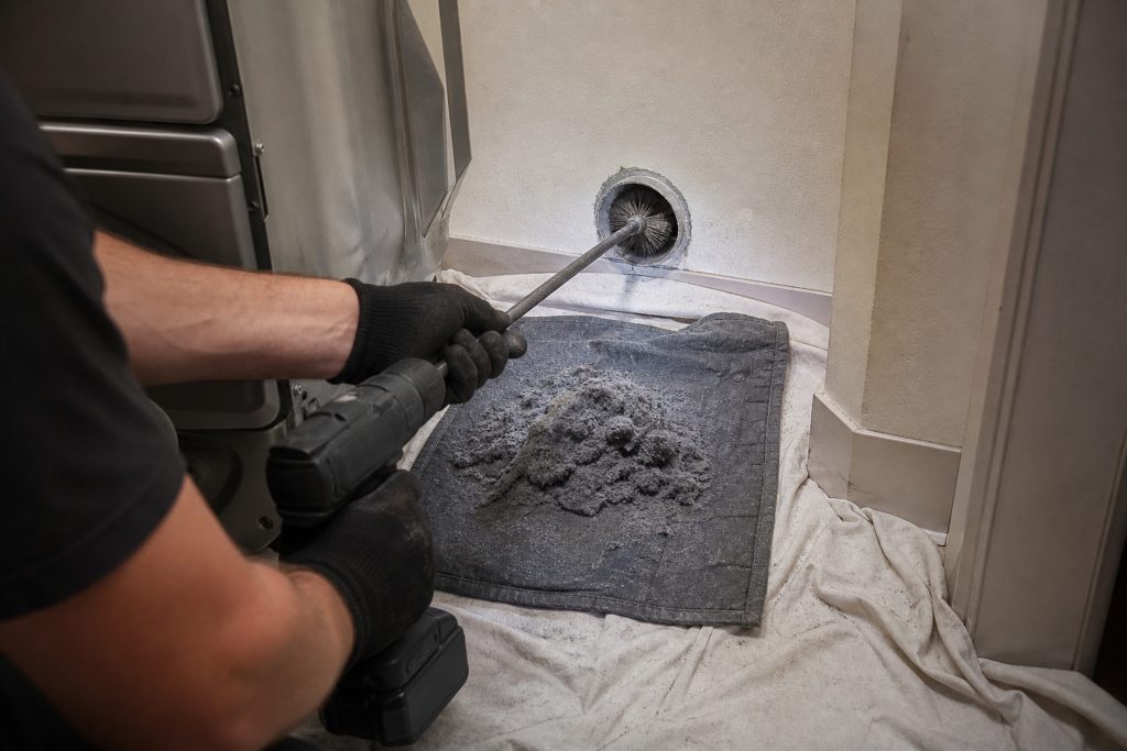 A rotary brush on a flexible rod driven by a cordless drill being fed into a wall duct behind a dryer that has been pulled forward, a grey mat of lint already pulled out onto a dust sheet on the floor.