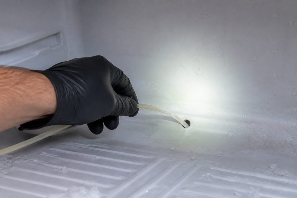 A gloved hand feeding a slim flexible tube into a small drain hole at the back of a freezer compartment, ice cleared away around it.